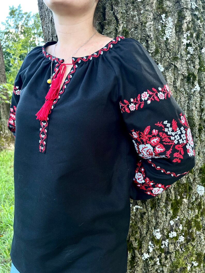 K&ouml;nnte beinhalten: Schwarze bestickte Bluse mit roten und wei&szlig;en Blumendesigns. Die Bluse hat ein Quasten-Detail am Ausschnitt.