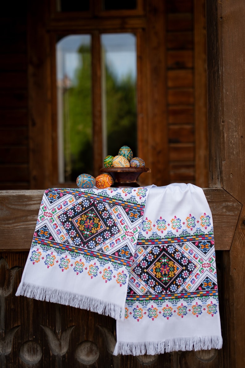 May include: A white embroidered table runner with a colorful geometric pattern. The runner is hanging on a wooden surface with a bowl of painted Easter eggs in the background.