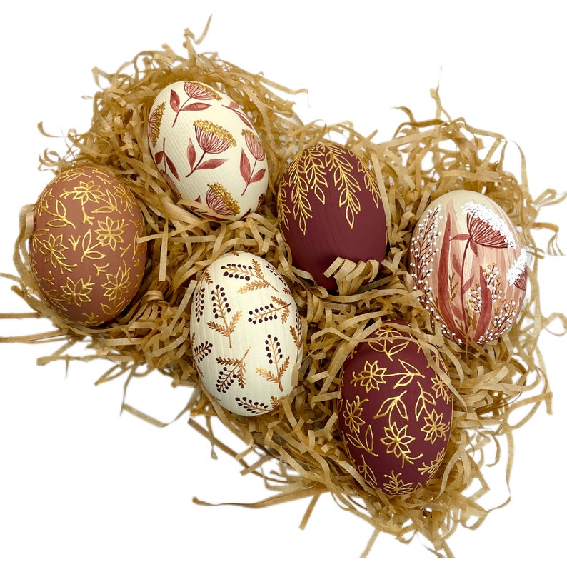 May include: Six hand-painted Easter eggs with gold and burgundy designs. The eggs are arranged in a nest of shredded paper.
