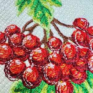 May include: Close-up of an embroidered design featuring a cluster of red berries with white highlights, green leaves, and brown stems. The embroidery is on a white, textured fabric, showcasing detailed needlework.