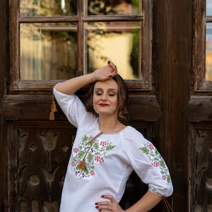 May include: A woman wearing a white embroidered blouse with green and red floral designs and brown tassels. She is standing in front of a wooden door with a window.