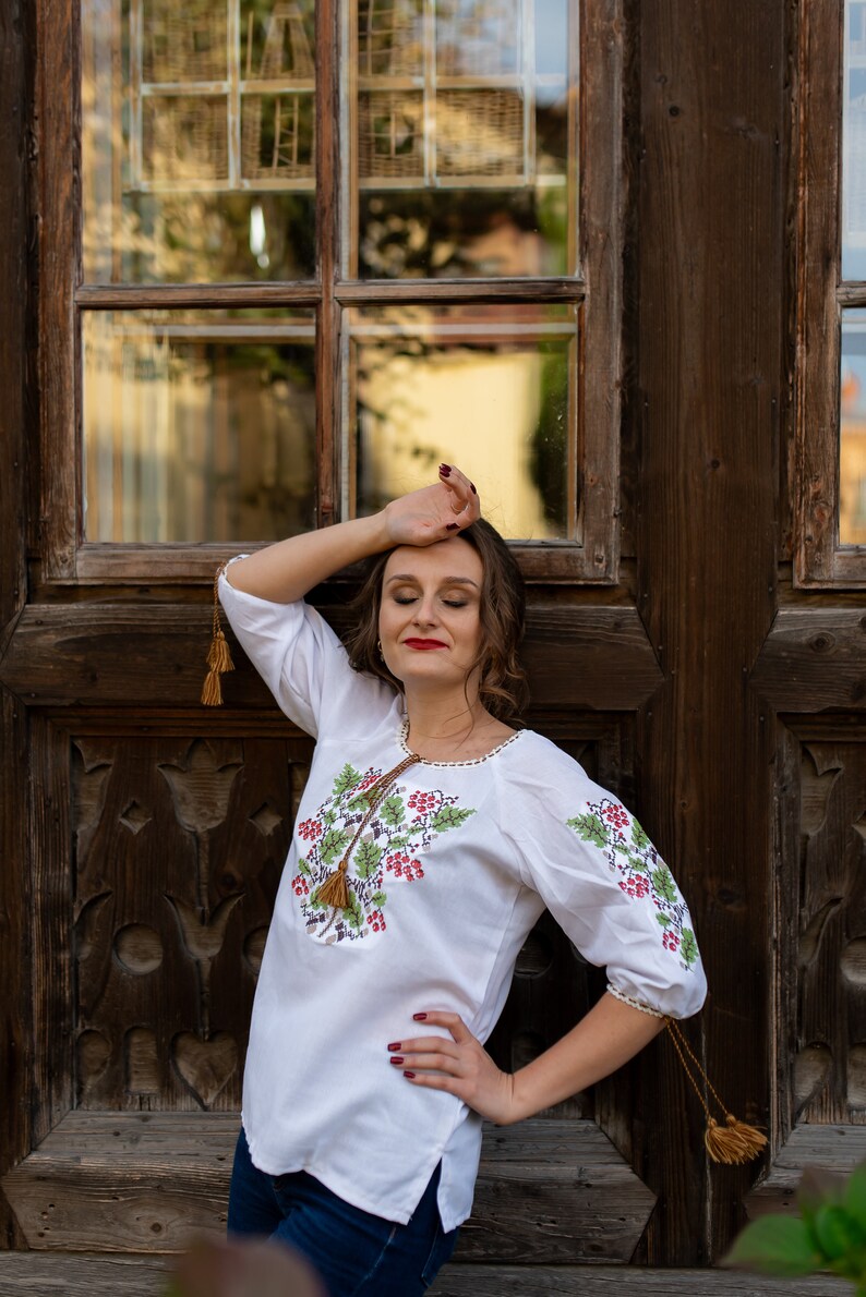 May include: A woman wearing a white embroidered blouse with green and red floral designs and brown tassels. She is standing in front of a wooden door with a window.