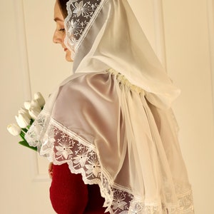May include: A sheer white veil with delicate floral lace trim. The veil is draped over the shoulders and head, creating a soft, elegant look. The floral lace is visible along the edges of the veil. The person is holding a bouquet of white tulips.