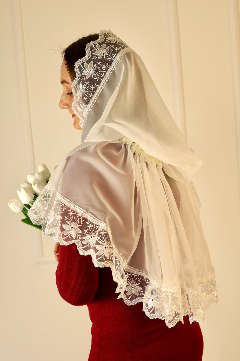 May include: A sheer white veil with delicate floral lace trim. The veil is draped over the shoulders and head, creating a soft, elegant look. The floral lace is visible along the edges of the veil. The person is holding a bouquet of white tulips.