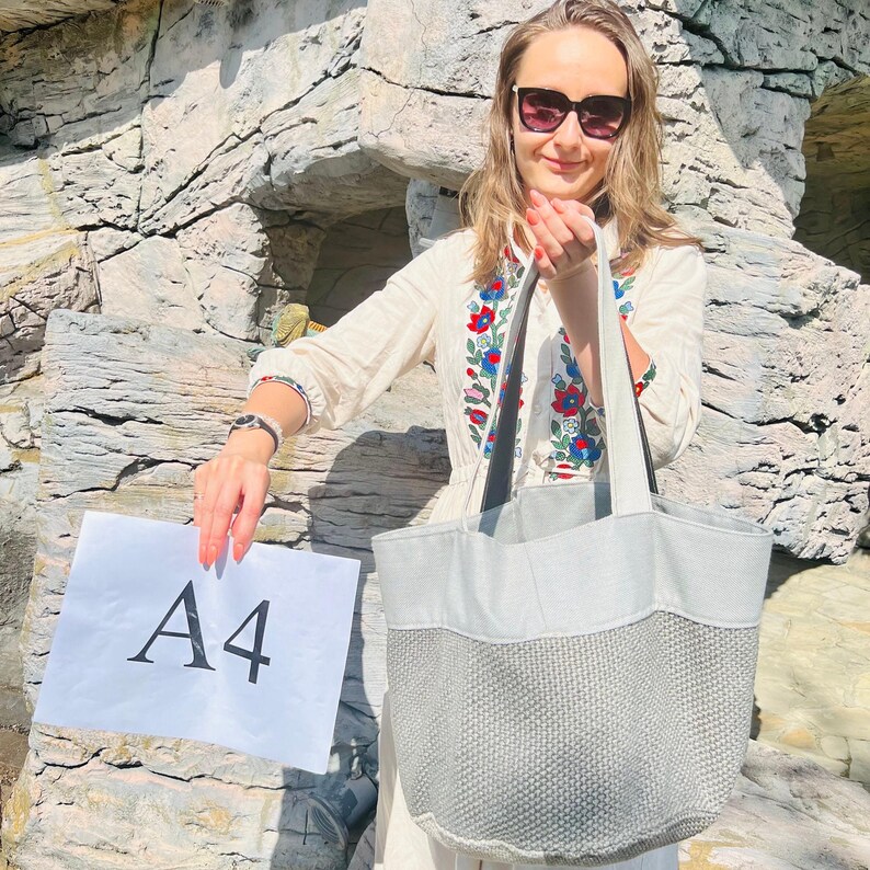K&ouml;nnte beinhalten: Eine hellgraue Tragetasche mit strukturiertem Unterteil und wei&szlig;en Griffen. Die Tasche wird vor einer Person gehalten, die eine Sonnenbrille und ein cremefarbenes, besticktes Oberteil tr&auml;gt. Ein wei&szlig;es A4-Papier wird in der anderen Hand der Person gehalten.