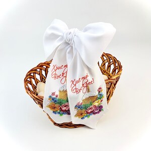 May include: A woven brown basket holds white eggs and two white fabric ribbons. The ribbons are embroidered with a cake design and red text. A large white bow is tied at the top of the ribbons.
