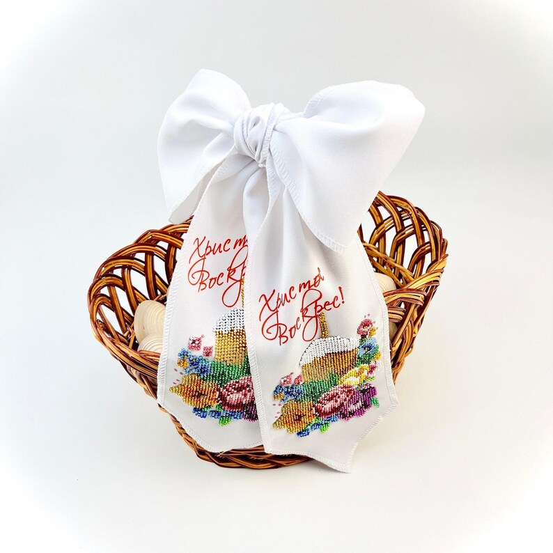May include: A woven brown basket holds white eggs and two white fabric ribbons. The ribbons are embroidered with a cake design and red text. A large white bow is tied at the top of the ribbons.
