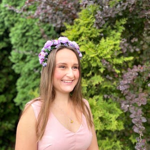 May include: A woman wearing a light pink dress and a floral crown with purple flowers. She is smiling, with long brown hair and a necklace with a yellow pendant. The background features green and purple foliage.