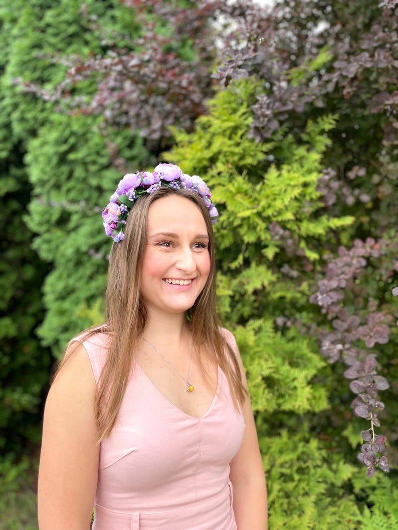 May include: A woman wearing a light pink dress and a floral crown with purple flowers. She is smiling, with long brown hair and a necklace with a yellow pendant. The background features green and purple foliage.