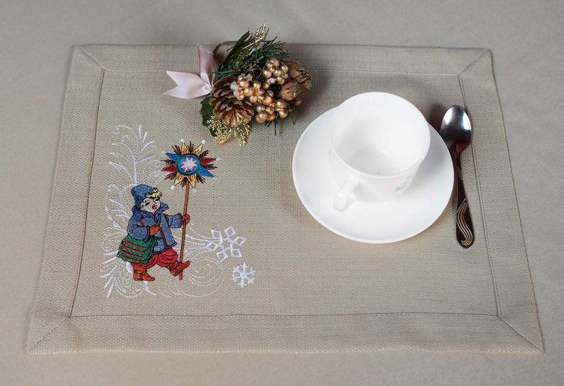 Op de afbeelding: Beige rechthoekige placemat met geborduurde wintersc&egrave;ne. Een kinderfiguur in een blauwe jas en rode broek houdt een stervormig object vast. Een witte kop en schotel, een lepel en een decoratief dennenappelarrangement zijn ook aanwezig.
