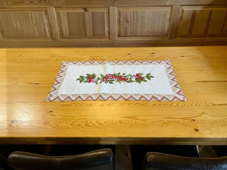 May include: A white crocheted table runner with a red berry and green leaf embroidery design on a wooden table.