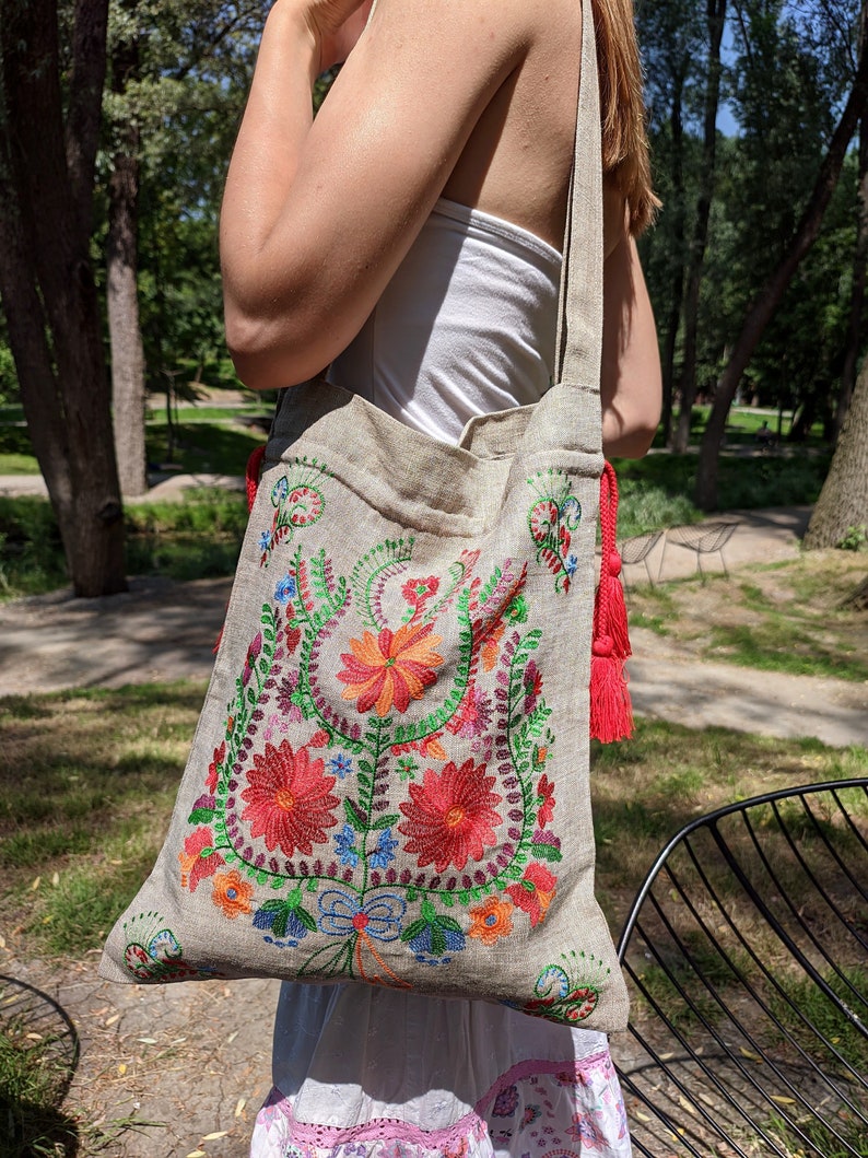 May include: A beige linen tote bag with a colorful floral embroidery design. The bag has a red tassel on the strap.