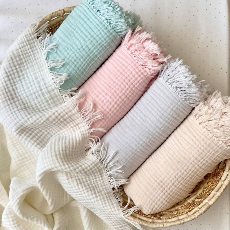 Peut inclure: Quatre couvertures pour b&eacute;b&eacute; douces et textur&eacute;es dans un panier. Les couvertures sont blanches, bleu clair, rose clair et beige. Chaque couverture a un bord frang&eacute;.