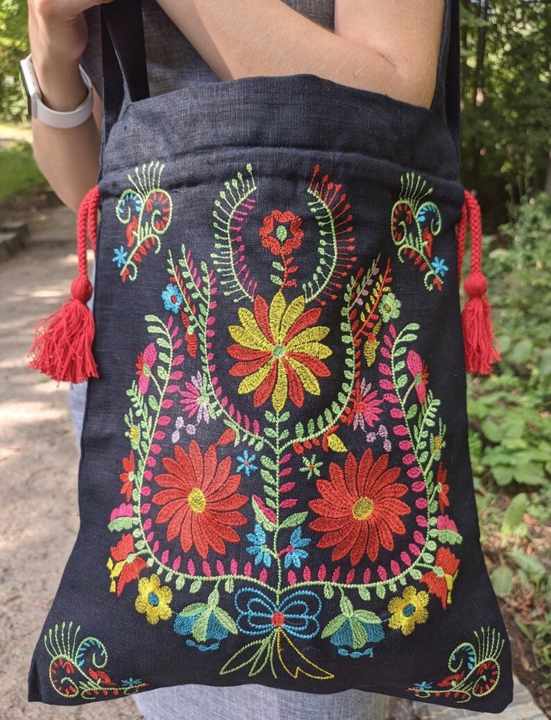 May include: Black tote bag with colorful embroidered floral design. The flowers are red, yellow, blue, green, and pink. The bag has red tassels on the sides.