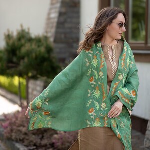 May include: A green shawl with floral embroidery. The shawl is draped over a person's shoulders and is worn with a brown top.