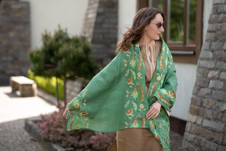 May include: A green shawl with floral embroidery. The shawl is draped over a person's shoulders and is worn with a brown top.