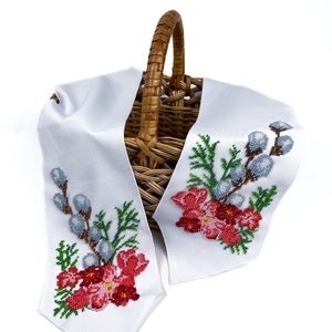 May include: A white fabric basket liner with embroidered floral designs featuring red flowers, green leaves, and blue willow branches. The liner is draped over a wicker basket.