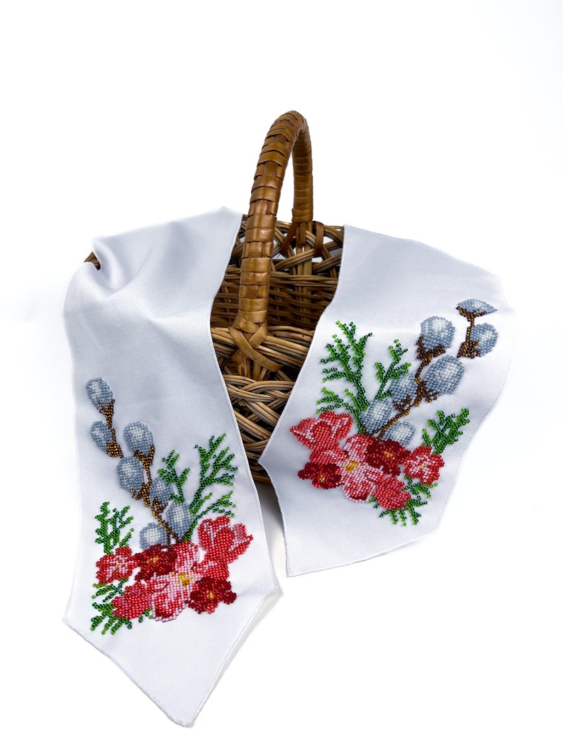 May include: A white fabric basket liner with embroidered floral designs featuring red flowers, green leaves, and blue willow branches. The liner is draped over a wicker basket.