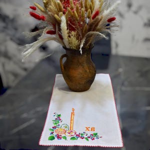 K&ouml;nnte beinhalten: Ein wei&szlig;er bestickter Tischl&auml;ufer mit rotem Rand. Die Stickerei zeigt eine Kerze, drei Ostereier und die kyrillischen Buchstaben "XB". Eine Vase mit Trockenblumen steht auf dem L&auml;ufer.