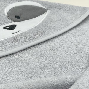 May include: A light grey hooded baby towel. The hood features a raccoon face design with grey, white, and black embroidered details. The towel is made of a soft, absorbent material, perfect for after bath time.