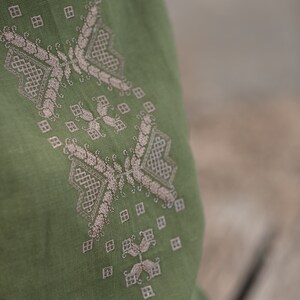 May include: A green linen top with a delicate embroidered design in a light beige colour. The embroidery features a geometric pattern with leaves and swirls.