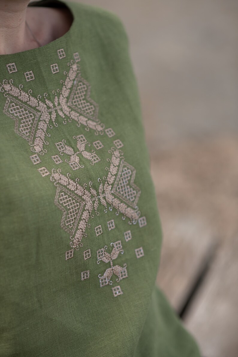 May include: A green linen top with a delicate embroidered design in a light beige colour. The embroidery features a geometric pattern with leaves and swirls.