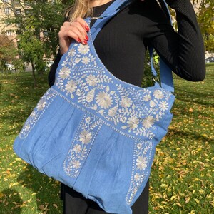Peut inclure: Un sac cabas en denim bleu clair avec des motifs floraux brod&eacute;s blancs. Le sac a une longue bandouli&egrave;re et une large ouverture.