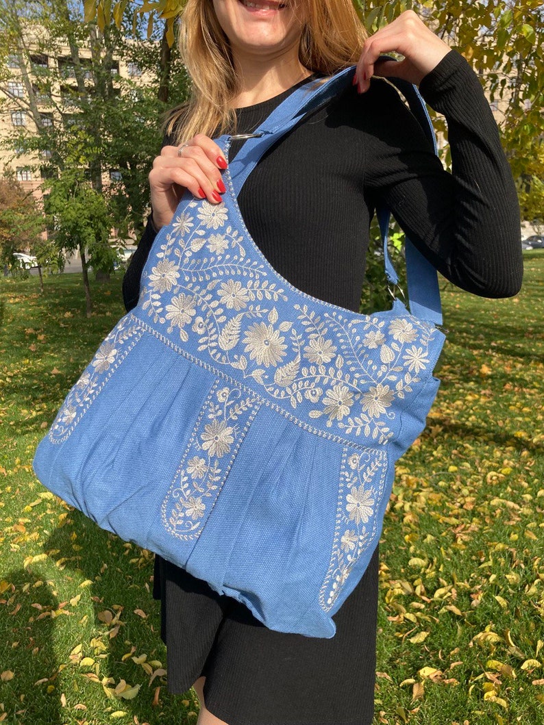 Peut inclure: Un sac cabas en denim bleu clair avec des motifs floraux brod&eacute;s blancs. Le sac a une longue bandouli&egrave;re et une large ouverture.