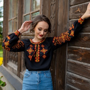 May include: A woman wearing a black embroidered blouse with red and yellow geometric patterns. The blouse has a long sleeve and a high neckline. She is standing in front of a wooden wall.