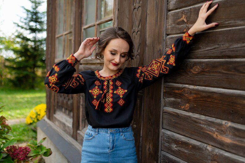 May include: A woman wearing a black embroidered blouse with red and yellow geometric patterns. The blouse has a long sleeve and a high neckline. She is standing in front of a wooden wall.