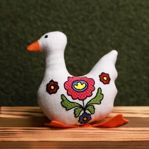 May include: A white fabric goose figurine with an orange beak and feet. The goose is decorated with embroidered flowers in red, pink, yellow, and blue, with green leaves. The figurine is sitting on a wooden surface.