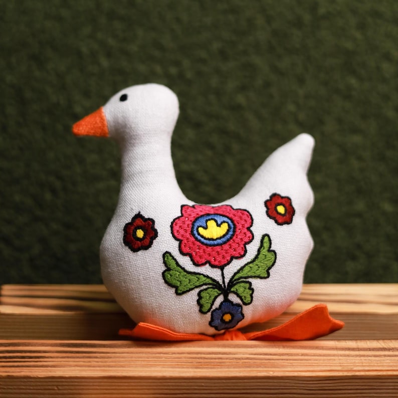May include: A white fabric goose figurine with an orange beak and feet. The goose is decorated with embroidered flowers in red, pink, yellow, and blue, with green leaves. The figurine is sitting on a wooden surface.
