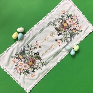 以下が含まれることがあります： 白いイースターテーブルランナー。鳥、巣、花の刺繍が施されています。ランナーは装飾的なボーダーと「Воскрес Христос」の文字が特徴です。パステルカラーのイースターエッグが、緑色の背景のランナーの周りに散らばっています。
