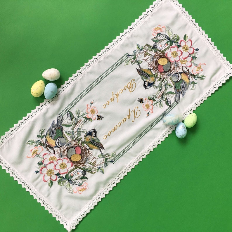 以下が含まれることがあります： 白いイースターテーブルランナー。鳥、巣、花の刺繍が施されています。ランナーは装飾的なボーダーと「Воскрес Христос」の文字が特徴です。パステルカラーのイースターエッグが、緑色の背景のランナーの周りに散らばっています。