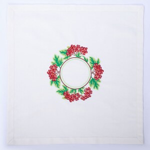 May include: White linen napkin with an embroidered wreath design. The wreath features red berries, green leaves, and a brown and green circular frame. The napkin has a simple, clean border.