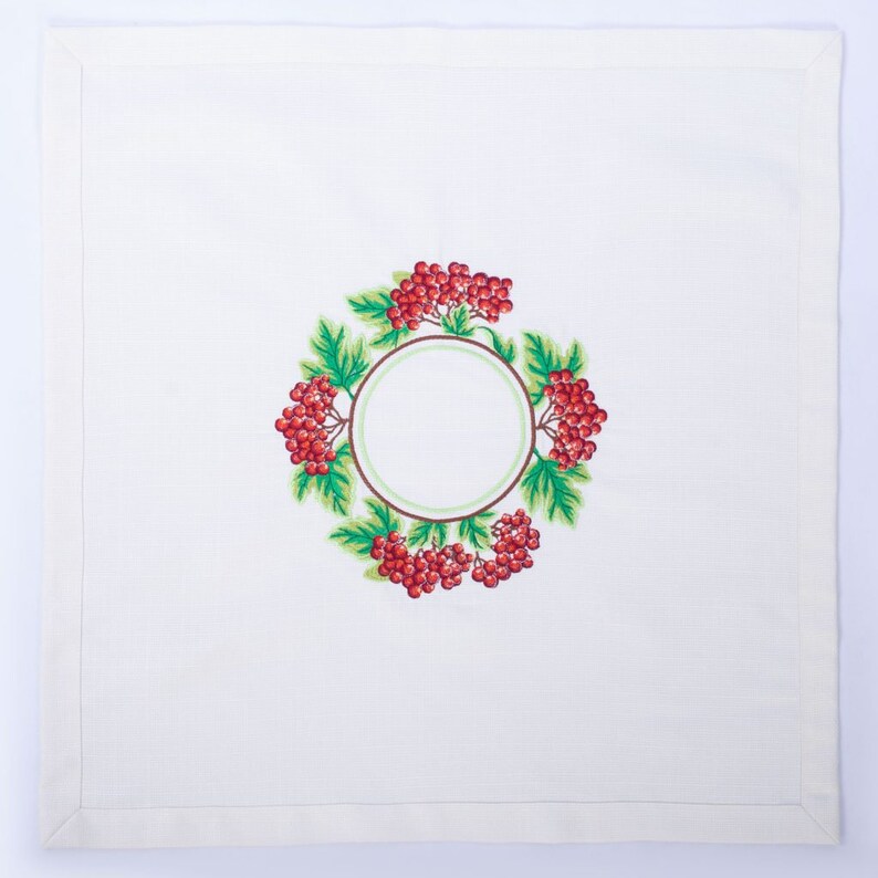 May include: White linen napkin with an embroidered wreath design. The wreath features red berries, green leaves, and a brown and green circular frame. The napkin has a simple, clean border.
