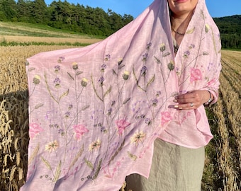 May include: A pale pink scarf featuring embroidered flowers in pink and purple, with green stems. The scarf is worn over the head and shoulders. The background shows a wheat field and a clear blue sky.