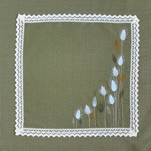 May include: Square olive green linen napkin with a white lace border. The corner features an embroidered floral design with white, light blue, and brown accents. The napkin is a decorative textile.