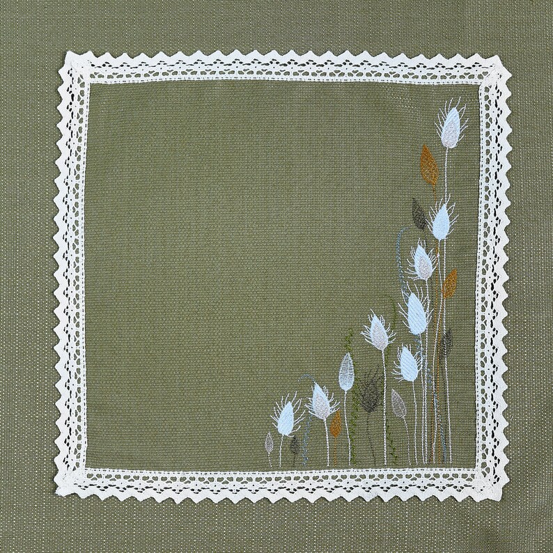 May include: Square olive green linen napkin with a white lace border. The corner features an embroidered floral design with white, light blue, and brown accents. The napkin is a decorative textile.