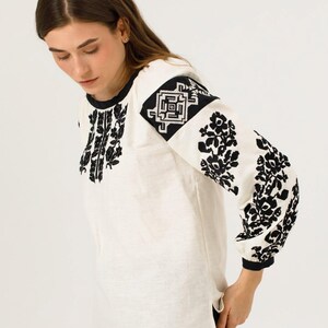 May include: A white blouse with long sleeves, adorned with detailed black floral embroidery on the sleeves and front. The blouse has a round neck and black cuffs. The design is in a traditional folk art style.