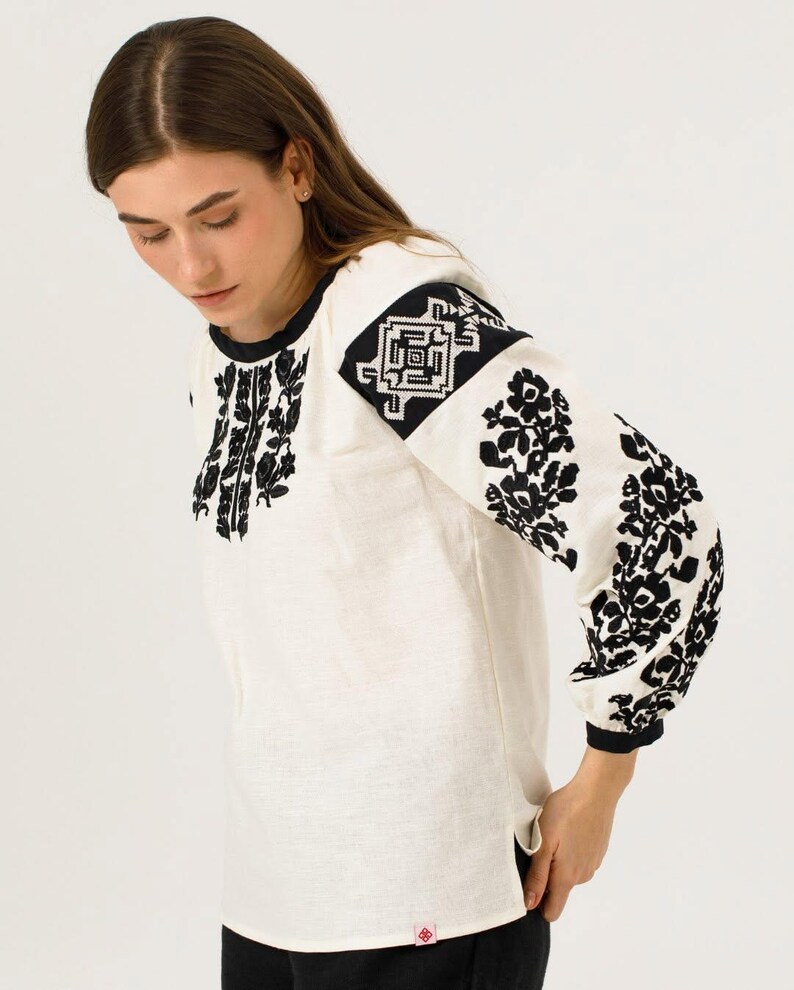 May include: A white blouse with long sleeves, adorned with detailed black floral embroidery on the sleeves and front. The blouse has a round neck and black cuffs. The design is in a traditional folk art style.