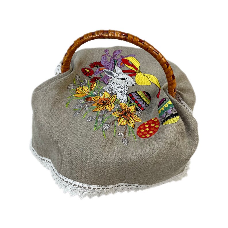 May include: A beige linen Easter basket cover with a white bunny, yellow daffodils, and colorful eggs embroidered on it. The cover has a brown wicker handle and a white lace trim.