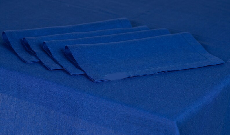 May include: Four folded, cobalt blue linen napkins arranged on a matching tablecloth. The napkins are neatly stacked, showcasing their crisp edges and textured surface. The deep blue color creates a cohesive and elegant table setting.