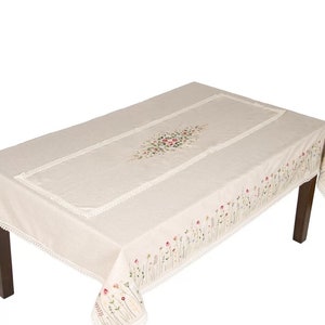 May include: A beige tablecloth with a white lace border and embroidered floral designs. The tablecloth is draped over a wooden table.