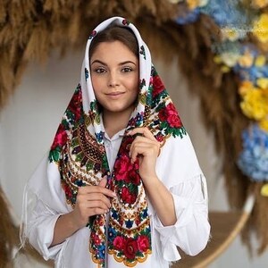May include: A white headscarf with a colorful floral pattern. The scarf is folded and tied around the head, with the floral pattern visible on the front and sides.