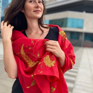 Puede incluir: Una mujer lleva una chal roja con bordados dorados. El chal tiene un dise&ntilde;o floral con p&aacute;jaros y hojas. La mujer lleva un vestido negro con estampado floral.