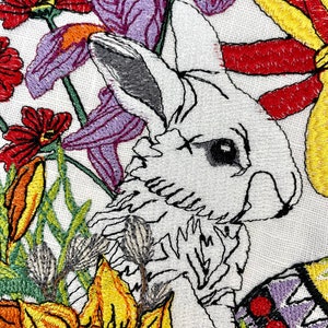 May include: Embroidered design featuring a white rabbit peeking out from behind a cluster of colorful flowers. The flowers include purple irises, yellow daffodils, and red blossoms. The rabbit's fur is detailed with white and gray stitching.