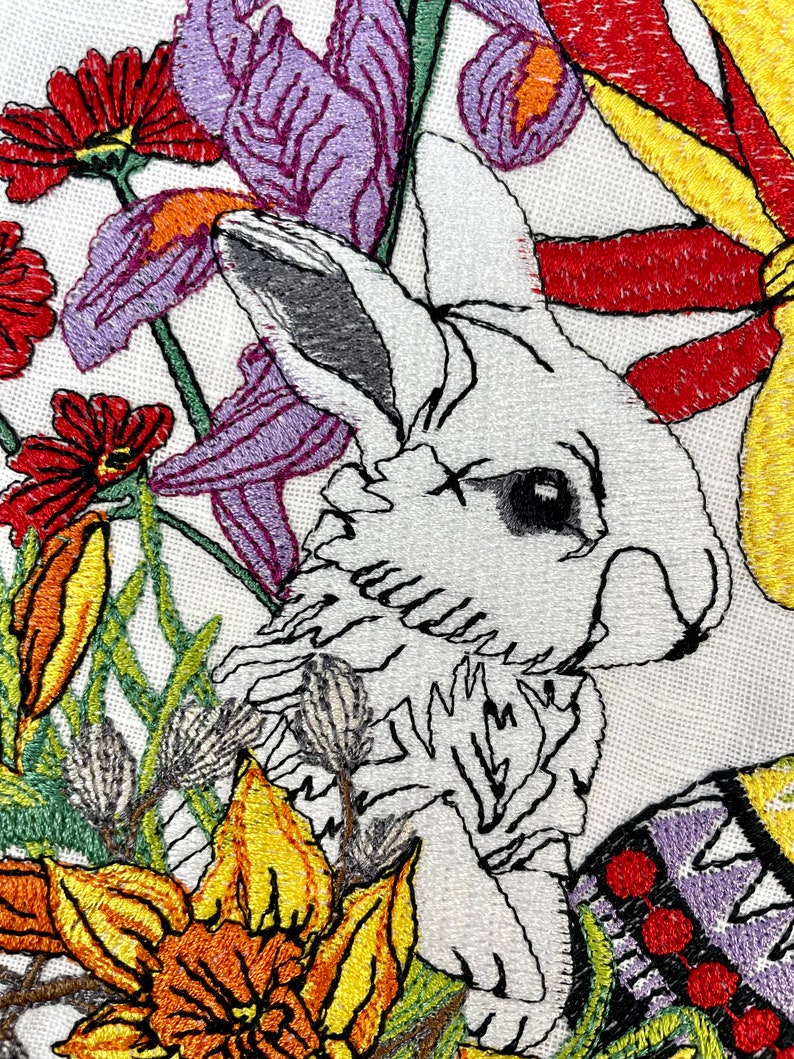 May include: Embroidered design featuring a white rabbit peeking out from behind a cluster of colorful flowers. The flowers include purple irises, yellow daffodils, and red blossoms. The rabbit's fur is detailed with white and gray stitching.