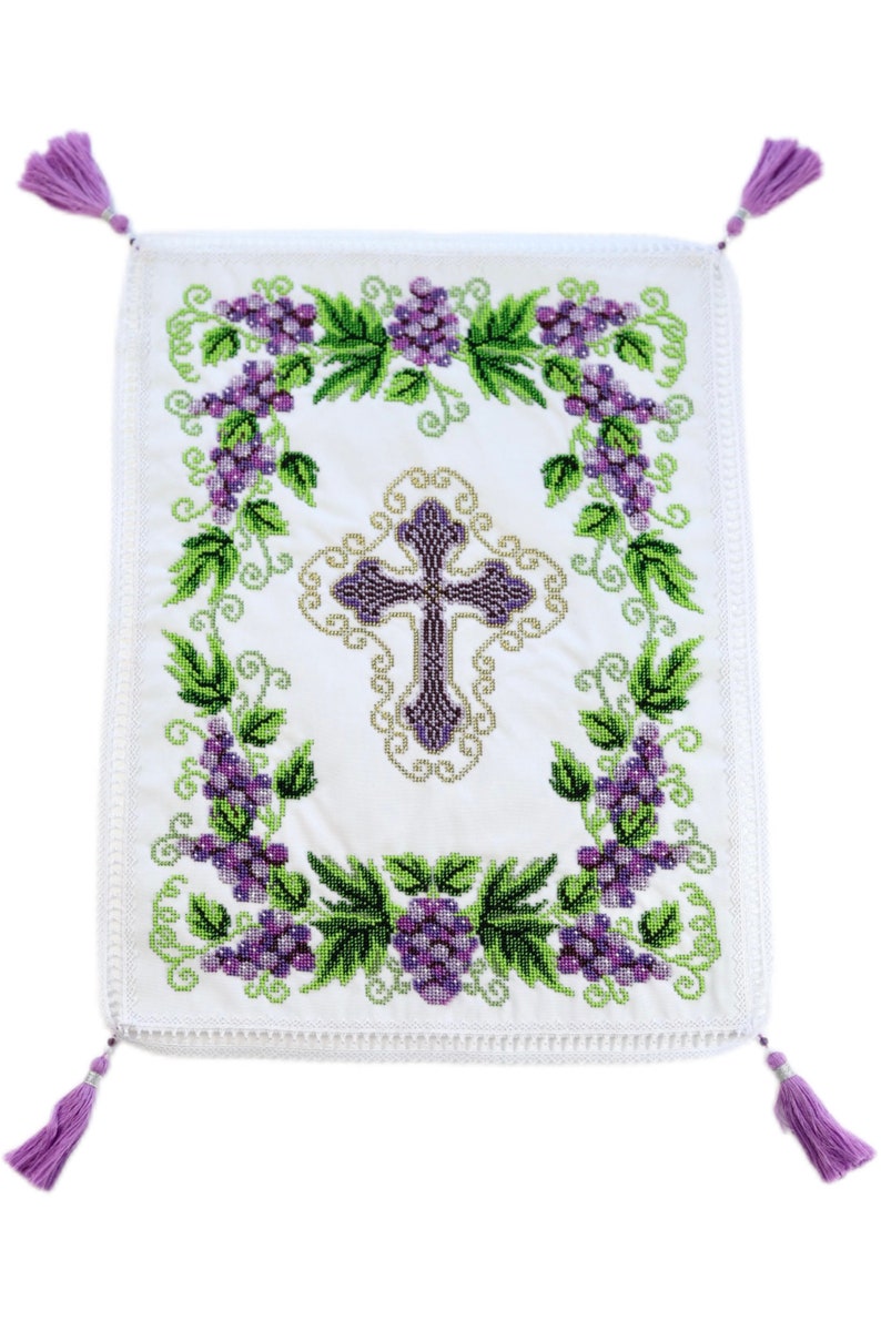 Puede incluir: Pa&ntilde;o rectangular blanco bordado con un dise&ntilde;o central de cruz morada. La cruz est&aacute; rodeada por un borde dorado y enmarcada por hojas verdes y racimos de uvas moradas. Cuatro borlas moradas est&aacute;n unidas a las esquinas.