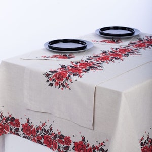 May include: A beige tablecloth and matching placemat set with a floral cross-stitch design in red and black. Two black plates sit on the table. The tablecloth is made of a natural fabric.